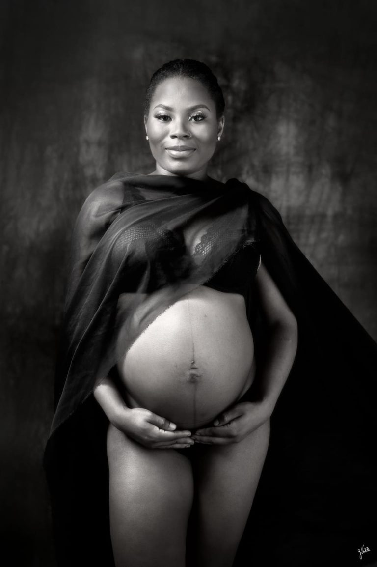 Photographe grossesse Marseille 13 Photographe de grossesse à Marseille – portrait en noir et blanc d’une femme enceinte debout en studio, posant face à l’objectif contre un fond noir texturé. Elle tient son ventre rond avec tendresse, enveloppée d’un voile noir qui souligne la douceur et l’intemporalité de la scène. Cette prise de vue en lumière intimiste, réalisée par un photographe professionnel spécialisé en maternité, évoque la transmission, l’élégance classique et la force tranquille de la grossesse. Un shooting photo grossesse raffiné, pensé pour créer des souvenirs inoubliables dans une ambiance studio